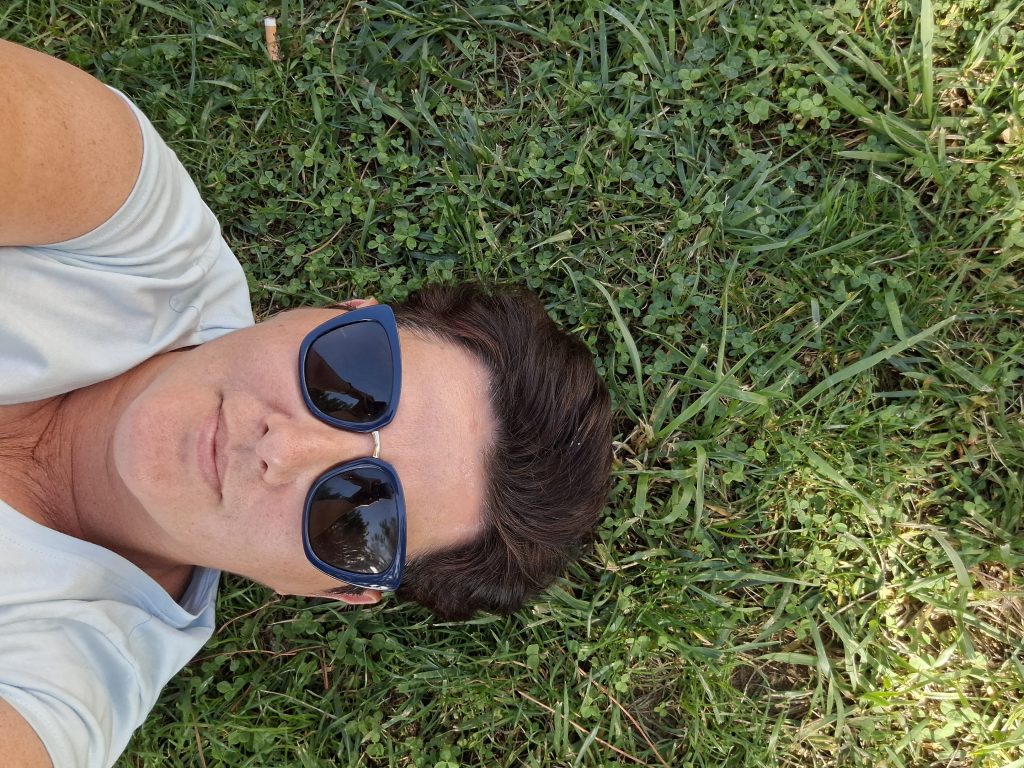 Zdjęcie typu selfie wykonane z góry, przedstawiające osobę o krótkich ciemnych włosach leżącą na zielonej trawie. Osoba ma na sobie okulary przeciwsłoneczne z ciemnymi oprawkami i jasny t-shirt.